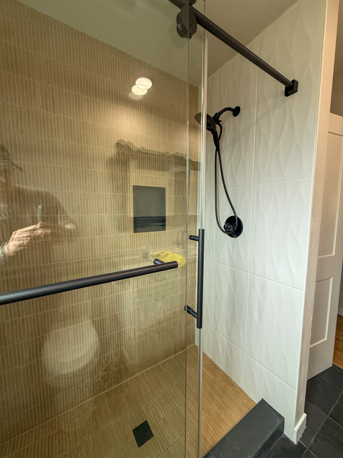 A contemporary shower area combining white textured wall tiles and beige vertical subway tiles, featuring a glass door and black hardware.