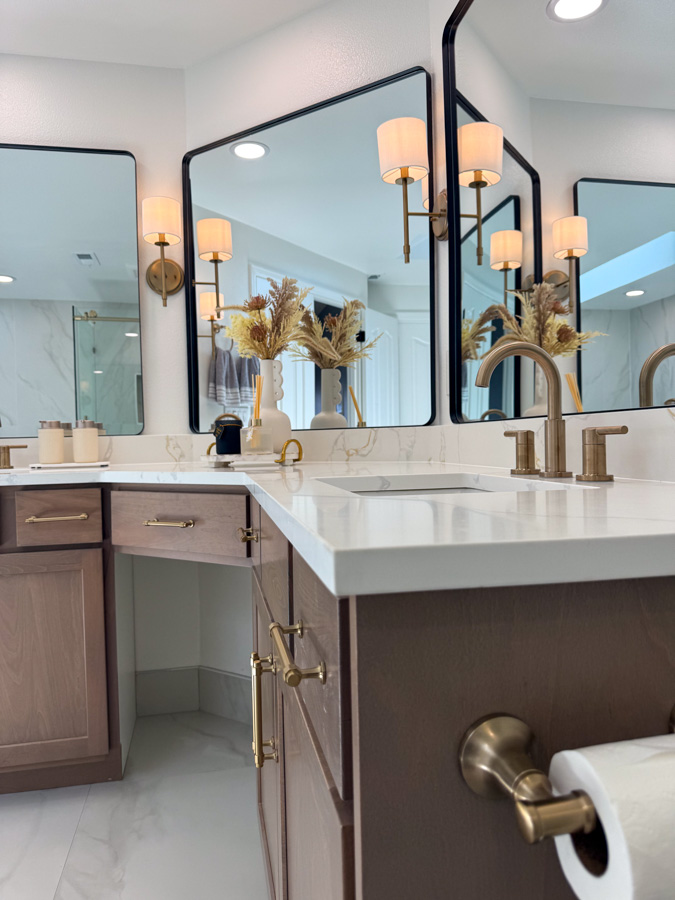 A well-lit bathroom vanity with a large mirror, brass fixtures, and decorative wall sconces, complemented by wooden cabinets.