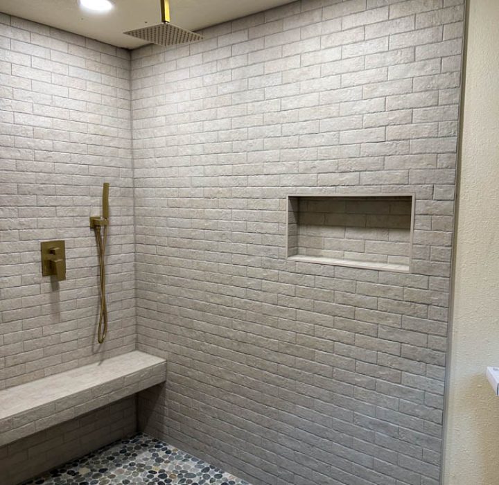 A minimalist shower area with light brick-tile walls, a pebble tile floor, and a built-in bench for added comfort.