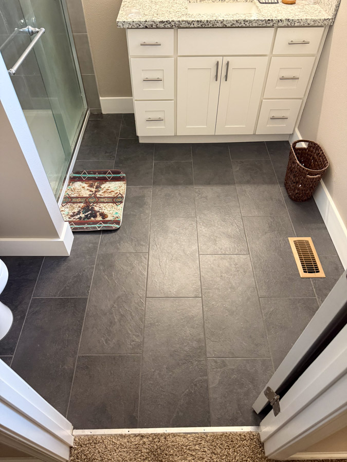 A stylish bathroom with dark floor tiles, white cabinets, and a patterned rug near the entrance.